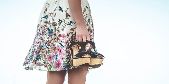 A woman in a floral dress holding a pair of high heeled shoes.