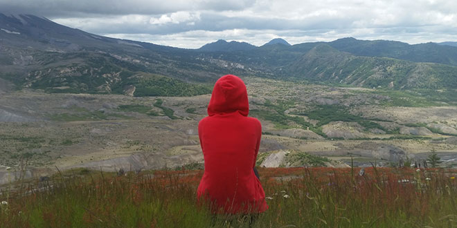 A woman in a red hoodie is standing in a field looking at mountains.