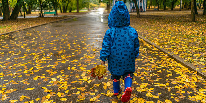 A child in a raincoat walking through a park with leaves.