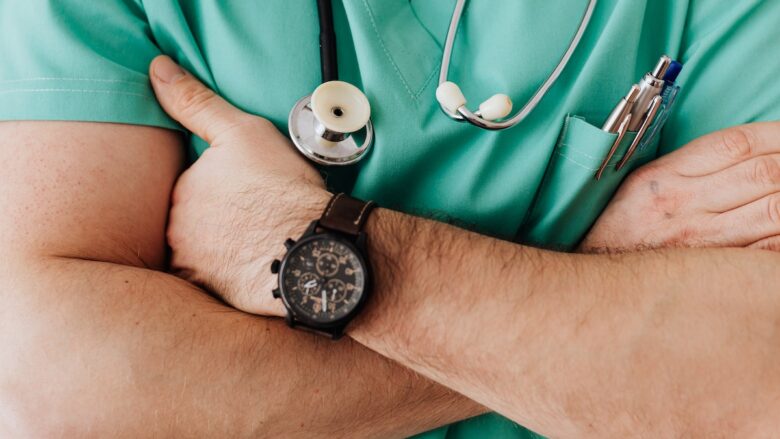 A male doctor with his arms crossed and a stethoscope and a watch on his wrist.